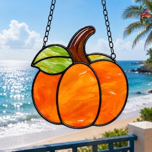 May include: A stained glass pumpkin suncatcher featuring vibrant orange glass segments, a brown glass stem, and a bright green glass leaf. The piece is framed in black metal solder and hangs from a black metal chain, showcasing a smooth texture.
