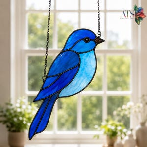 Puede incluir: Atrapasoles de vidriera con forma de pájaro azul, suspendido por una cadena negra. El pájaro presenta diferentes tonos de azul y un pico marrón. El fondo muestra una ventana con vegetación. Diseño de estilo mosaico.