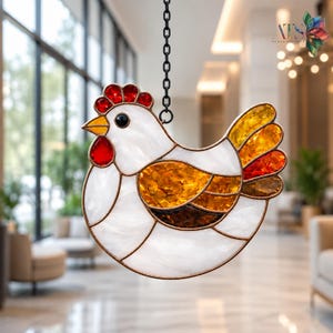 Könnte beinhalten: Ein Hühner-Sonnenfänger aus Buntglas mit weißen, bernsteinfarbenen und roten Glassegmenten, die durch dunkle Metalllötlinien verbunden sind. Das Huhn hat einen roten Kamm und Kehllappen, einen gelben Schnabel und ein schwarzes Auge an einer Kette.