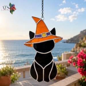 May include: A stained glass suncatcher shaped like a black cat wearing an orange witch hat with a purple band and a yellow buckle. The piece is constructed with dark glass segments joined by silver metal solder lines and hangs from a black chain.