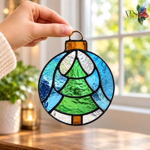 May include: A round stained glass ornament featuring a central green Christmas tree design surrounded by textured blue and clear glass panels. The ornament has a dark metal frame and a small amber-colored glass cap with a metal hanging loop.