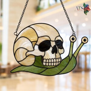 May include: Stained glass skull snail with a cream-coloured skull, black eye sockets, and a white-toothed jaw. The snail's body is green, and the eyes are black and white. The piece is suspended by a black chain. The text "ATS GLASS PATTERN" is visible.