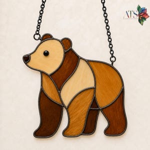 Può includere: Un acchiappasole in vetro colorato a forma di orso, realizzato con pezzi di vetro in varie tonalità di marrone e beige uniti da linee di saldatura in metallo scuro. L'orso ha un occhio in vetro nero ed è appeso a una catena di metallo nero.