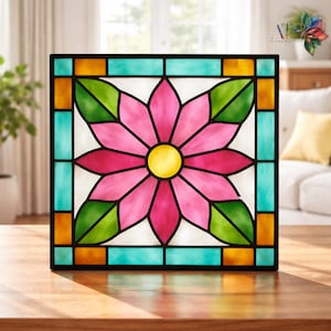 May include: Square stained glass panel featuring a pink flower with a yellow center and green leaves. The panel is framed in black with turquoise and orange accents. The panel is sitting on a wooden surface.