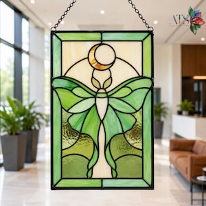 Pode incluir: Um apanhador de sol retangular em vitral apresentando uma mariposa verde central com asas alongadas sob uma lua crescente dourada. A peça é emoldurada em metal preto e suspensa por uma corrente preta, exibindo vários tons de vidro verde.