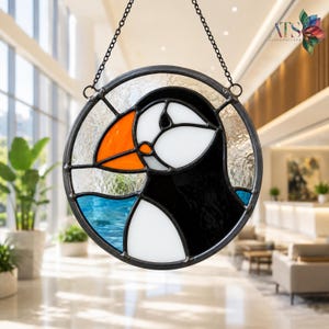 May include: A circular stained glass suncatcher featuring a puffin design with a bright orange beak, white face, and black body. The background consists of textured clear glass and a blue section at the bottom, all held together by dark metal cames.