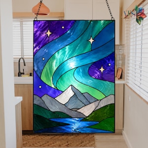 Op de afbeelding: Gebrandschilderd glaspaneel dat een nachthemel met het noorderlicht uitbeeldt. Het kunstwerk toont wervelende blauwe, groene en paarse tinten, met witte bergtoppen en een blauwe rivier aan de onderkant. Bevat drie sterren.