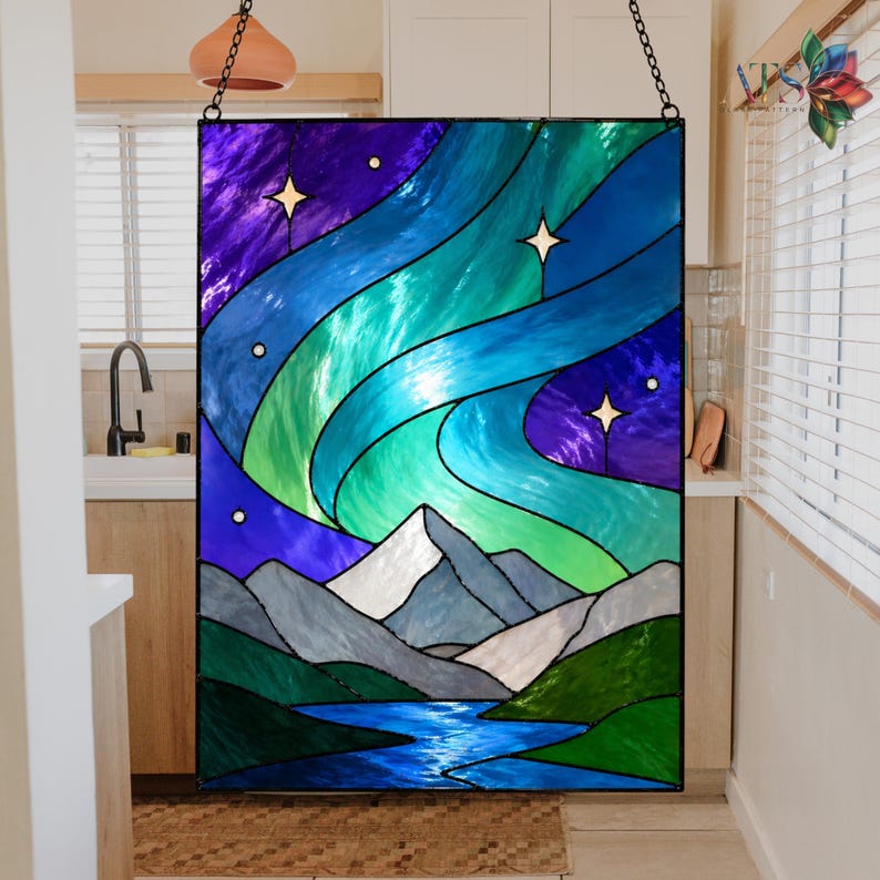 Op de afbeelding: Gebrandschilderd glaspaneel dat een nachthemel met het noorderlicht uitbeeldt. Het kunstwerk toont wervelende blauwe, groene en paarse tinten, met witte bergtoppen en een blauwe rivier aan de onderkant. Bevat drie sterren.