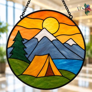 May include: A circular stained glass suncatcher featuring a landscape scene with a yellow sun, white-capped blue mountains, a green pine tree, a blue lake, and an orange tent on a green grassy foreground, all enclosed in a black metal frame.