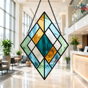 Op de afbeelding: Een ruitvormige glas-in-lood zonnevanger met een geometrisch patroon van getextureerde glaspanelen in tinten blauwgroen, lichtblauw, amber en helder wit. Het stuk is omlijst in zwart metaal en hangt aan een zwarte metalen ketting.