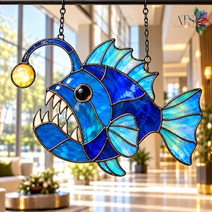 Peut inclure: Un attrape-soleil en vitrail en forme de baudroie avec des panneaux de verre bleu profond et turquoise, un leurre jaune lumineux et des dents blanches pointues. L'objet est suspendu par des chaînes en métal noir et porte le texte ATS Glass Pattern.