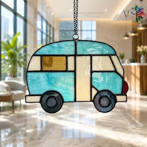 May include: A stained glass suncatcher shaped like a vintage camper van. The piece features light blue, cream, and amber glass panels held together by dark metal solder lines. It hangs from a black metal chain and includes a small red glass detail.