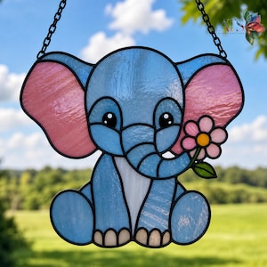 May include: A stained glass elephant suncatcher in shades of blue and pink, holding a flower. The elephant has large ears and is outlined in black. It hangs from a black chain. The background is a blurred view of a green field and blue sky.