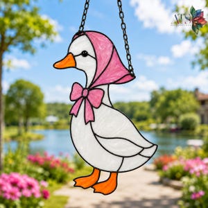 May include: A stained glass suncatcher shaped like a white duck wearing a pink headscarf and a matching pink bow. The duck has an orange beak and orange webbed feet, with black solder lines defining the edges of the white, pink, and orange glass.