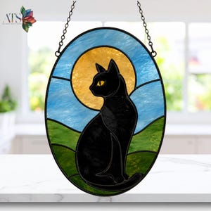 May include: An oval stained glass suncatcher featuring a black cat silhouette centered against a large, circular golden yellow moon. The background depicts a light blue sky above rolling green hills, all framed by a thin black metal border.