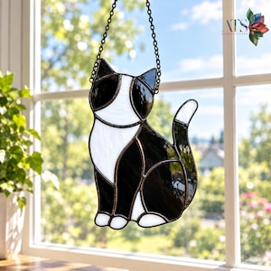 May include: A stained glass suncatcher shaped like a sitting cat with black and white glass segments joined by dark metal solder lines. The cat has a white chest and face markings with black ears and body, suspended by a black metal chain.