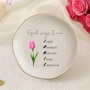 May include: A round white ceramic trinket dish with a gold-toned rim. The center features a pink tulip illustration and the text: God says I am J-oyful, A-bundant, M-ade new, I-mage, E-mpowered, spelling out the name JAMIE in a vertical black font.