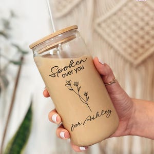 May include: A clear glass tumbler with a bamboo lid and a straw, filled with a light brown beverage. The tumbler has the words "Spoken over you" and "for Ashley" in black script, along with a floral design.