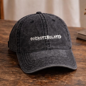 May include: A dark gray baseball cap with the word "OVERSTIMULATED" embroidered in white across the front. The cap has a curved brim and a fabric strap closure on the back. The hat is sitting on a wooden surface.