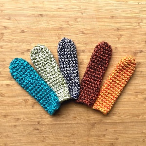 Puede incluir: Cinco fundas tejidas a mano en varios colores: turquesa, verde claro, azul marino, rojo oscuro y naranja. Las fundas están dispuestas sobre una superficie de madera, mostrando su apariencia texturizada y hecha a mano. Estos artículos son ideales para proteger objetos.