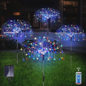 Op de afbeelding: Vier op zonne-energie werkende decoratieve buitenlampen met kleurrijke LED-verlichting in een vuurwerkachtig ontwerp. De lampen staan op metalen palen en staan in een grasveld. Een zonnepaneel en afstandsbediening zijn ook zichtbaar.