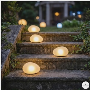 Puede incluir: Escena al aire libre con luces brillantes en forma de guijarro colocadas en escalones de piedra. Las luces, que emiten un brillo cálido, están dispuestas a lo largo de los escalones, creando un ambiente suave. La escena sugiere un entorno exterior tranquilo.