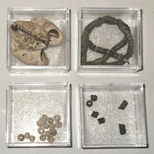 Könnte beinhalten: Vier quadratische, durchsichtige Schaukästen mit verschiedenen Objekten. Ein Kasten enthält drei cremefarbene Kaurimuscheln. Ein weiterer enthält eine dunkelgrüne, gewickelte Drahtkette. Der dritte enthält kleine runde beige Perlen und der vierte fünf dunkle Metallspiralen.