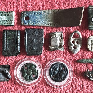 Könnte beinhalten: Eine Sammlung verschiedener antiker Metallartefakte, darunter rechteckige Platten mit geprägten Mustern, kreisförmige Medaillons, kleine Schnallen und Fragmente von Bronzewerkzeugen. Die Stücke weisen verwitterte Texturen, dunkle Patinas und geometrische Designs auf.