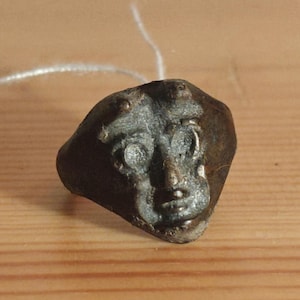Puede incluir: Un pequeño anillo antiguo de bronce con un rostro detallado. El rostro presenta ojos, nariz y boca prominentes, con una apariencia ligeramente desgastada. El anillo está sobre un fondo de madera.