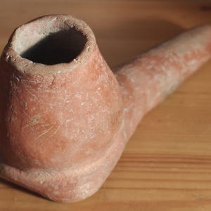 Puede incluir: Una pipa de arcilla color terracota con un hornillo redondeado y un tallo largo y cónico. La superficie de la pipa tiene un acabado mate y texturizado con imperfecciones naturales visibles y una apariencia porosa de tono marrón rojizo.