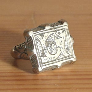 Puede incluir: Un anillo de plata con una cara cuadrada con un diseño grabado. La cara del anillo tiene un borde adornado y un diseño central con una inicial. El anillo está sobre una superficie de madera.