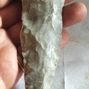 May include: A light gray, elongated stone tool with a pointed tip and a wide, flat base. The surface features distinct, irregular flake scars across its entire length, indicating a knapped construction with sharp, jagged edges along the sides.