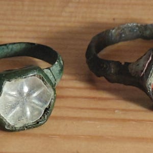 Puede incluir: Dos anillos antiguos con engastes ornamentados. Uno tiene una piedra transparente en forma de estrella, el otro una piedra roja y transparente. Los anillos se exhiben sobre una superficie de madera, mostrando su aspecto envejecido y desgastado.