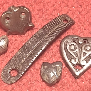 Peut inclure: Un ensemble de cinq objets métalliques comprenant une longue pièce rectangulaire étroite avec des lignes gravées en diagonale et un trou circulaire, une pièce en forme de cœur ajourée, deux petits charms en forme de cœur et une perle texturée.