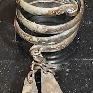 Puede incluir: Un anillo plateado con un diseño en espiral. El anillo tiene un dije colgante con dos pequeños colgantes rectangulares. Cada colgante tiene un diseño de estrella. El anillo está sobre una superficie oscura.