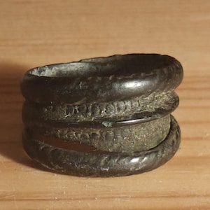 Puede incluir: Un anillo desgastado de color bronce con un diseño apilado. El anillo presenta tres bandas, con textura visible y signos de edad. Las bandas son ligeramente irregulares. El anillo está sobre una superficie de madera.