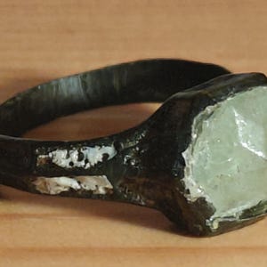 Puede incluir: Un anillo antiguo y oscuro con una piedra hexagonal de color verde pálido. La banda del anillo es oscura y parece ser de metal. La piedra está engastada en un engaste de bisel. El anillo se muestra sobre una superficie de madera.