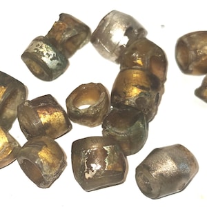 Può includere: Una collezione di piccole perline a forma di barilotto con un corpo in vetro traslucido color marrone grigiastro e inserti in lamina metallica dorata o argentata. Ogni perlina presenta un foro centrale per l'infilatura e superfici irregolari.
