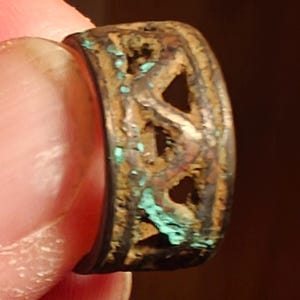 Pode incluir: Um close-up de um anel desgastado, cor de bronze, com um design vazado. O anel tem uma forma retangular com recortes triangulares e áreas de oxidação turquesa. O anel é segurado entre os dedos.