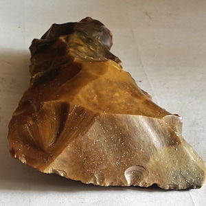 May include: A jagged, elongated piece of brown flint stone with a glossy, translucent surface. The object features sharp, irregular edges and a textured, multi-faceted appearance created by conchoidal fractures across its entire surface.