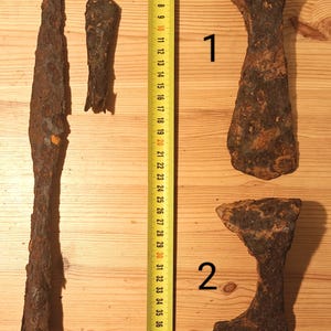 Può includere: Quattro antichi utensili in metallo arrugginito sono esposti su una superficie di legno accanto a un metro a nastro giallo. Gli utensili includono due teste d'ascia e altri due oggetti allungati e appuntiti. Il metro a nastro mostra centimetri.
