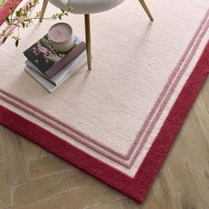 May include: A rectangular rug with a light beige center and a deep red border. Two lighter pink stripes separate the border from the center. A stack of books and a candle sit on the rug near a chair.
