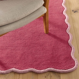 May include: A raspberry-colored rug with a scalloped edge and a light pink border. The rug is placed on a wooden floor, with a portion of a chair visible in the upper left corner. The rug's texture appears soft and plush.