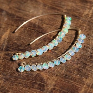 May include: A pair of curved gold wire earrings featuring a row of small, round, iridescent white opal beads. Each earring is constructed with a thin, arched metal wire that holds the beads in place, creating a delicate, crescent-shaped design.