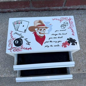 May include: White, two-drawer box with a poker-themed design. The top features a skull wearing a cowboy hat and a red bandana, smoking a cigarette. Text includes "feeling lucky" and "you cannot change the hand you are dealt, just the way you play the cards."