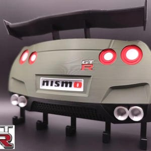 Può includere: Modello stampato in 3D della parte posteriore di un'auto, con spoiler, fanali posteriori e tubi di scarico. Il modello è verde oliva con dettagli rossi e i loghi "NISMO" e "GT-R". Include ganci.