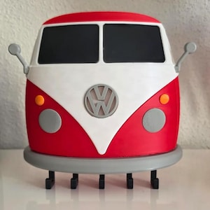 Könnte beinhalten: Ein rot-weißer Volkswagen Bus Schlüsselhalter zur Wandmontage mit einem zentralen VW-Logo, zwei schwarzen Windschutzscheiben, zwei grauen Scheinwerfern, zwei orangefarbenen Blinkern, zwei Seitenspiegeln und fünf schwarzen Haken am grauen Stoßfänger.