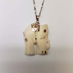 May include: A gold chain necklace with a carved ivory elephant pendant. The elephant has a gold saddle and a red gemstone on its trunk.