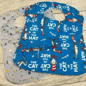 May include: Two baby bibs with snap closures. One bib is bright blue with a repeating pattern of The Cat in the Hat characters, red and white striped hats, and the text The Cat in the Hat. The second bib is light gray with a pattern of road signs.