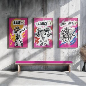 May include: Three framed art prints featuring zodiac signs: Leo, Aries, and Sagittarius. Each print has a pink background with a gold frame. The Leo print shows a cowboy, Aries features a ram skull, and Sagittarius depicts an archer on horseback.
